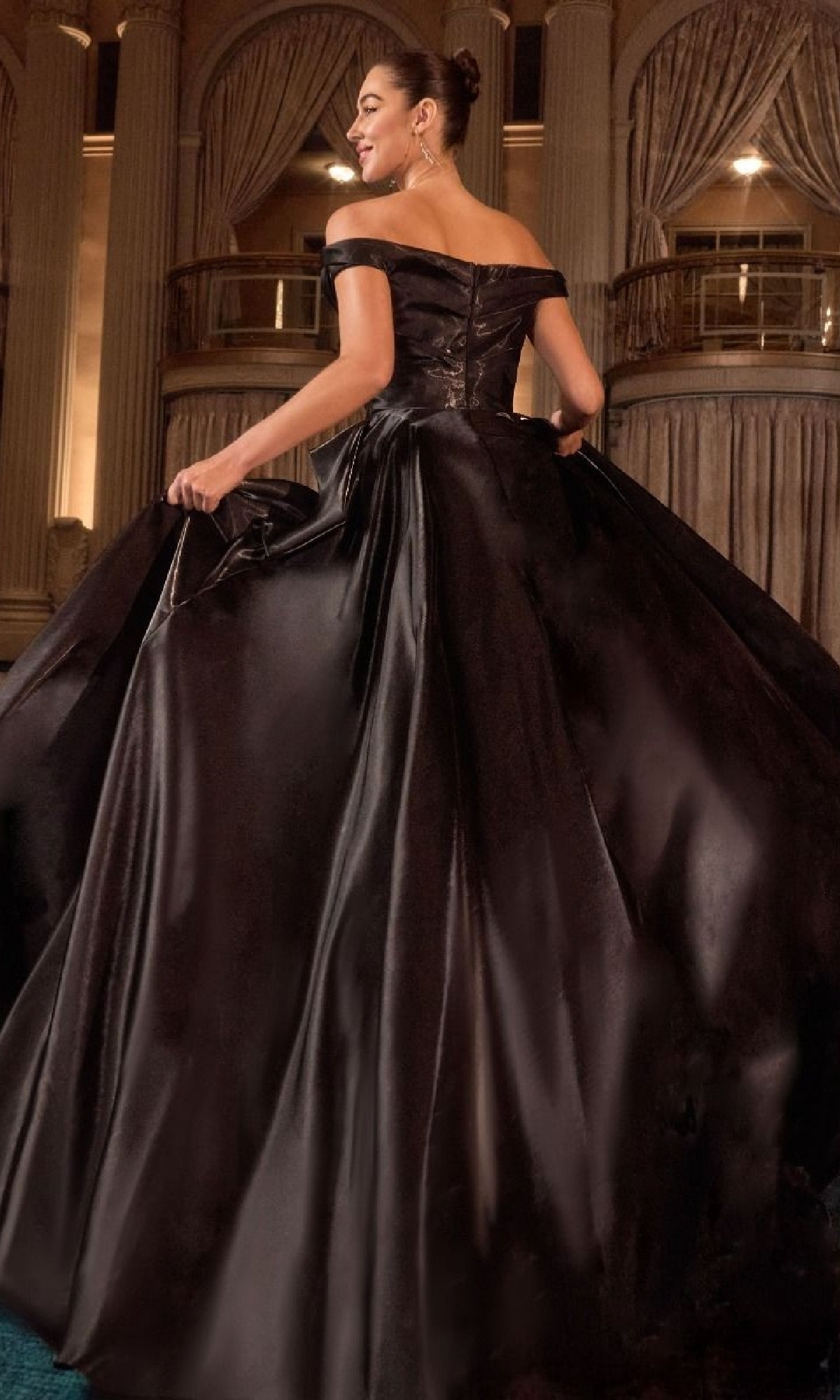 A woman stands in an ornate hall, smiling over her shoulder as she holds the skirt of the Ladivine J885 long prom dress.